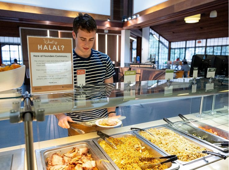 A young man in a striped shirt serves himself food from a cafeteria buffet with trays of noodles, rice, and meat, under a sign explaining halal food. The dining area is modern with large windows.