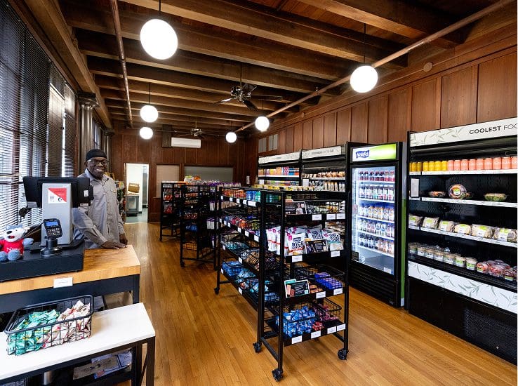 A small convenience store with wooden floors and exposed beams, featuring shelves stocked with snacks and drinks. A smiling employee stands behind the counter on the left. Bright lights hang from the ceiling.
