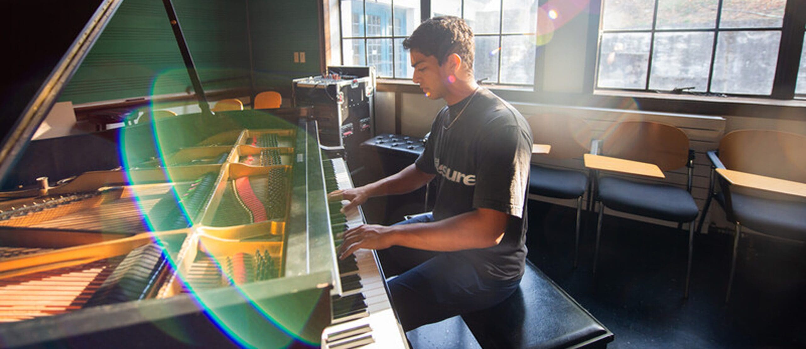 A person plays a grand piano in a sunlit room with empty chairs and a window in the background, colorful light flares visible in the foreground.