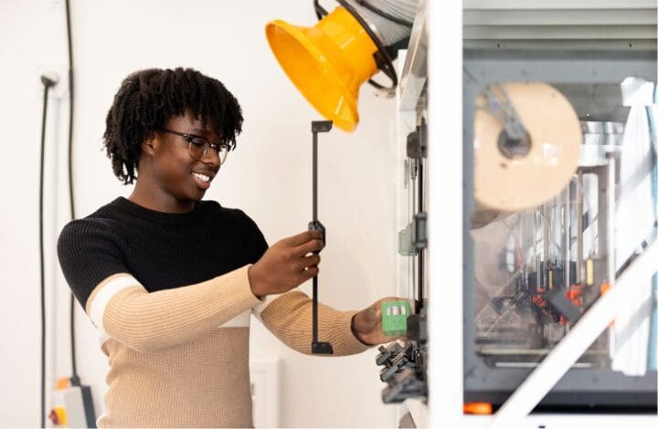 A person with glasses and curly hair, wearing a black and beige sweater, smiles while working with a 3D printer. They are holding a tool beside the machine, with a yellow lamp positioned above.