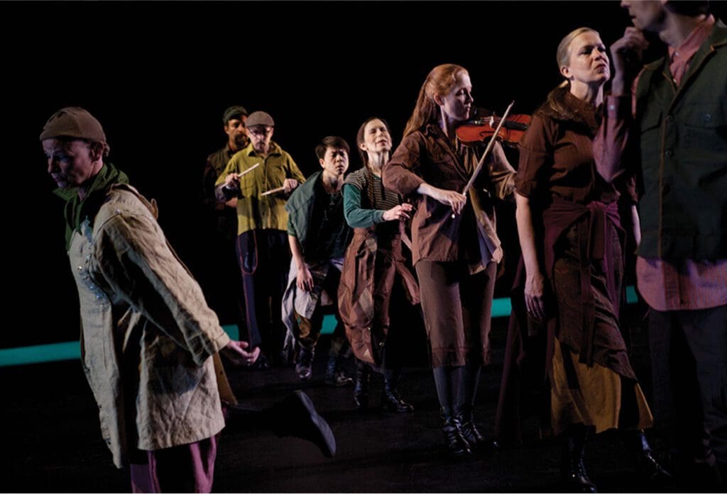 A group of performers in earth-toned costumes stands on a dark stage; one plays a violin, others appear to sing or act, some in mid-motion, suggesting a theatrical or musical performance.