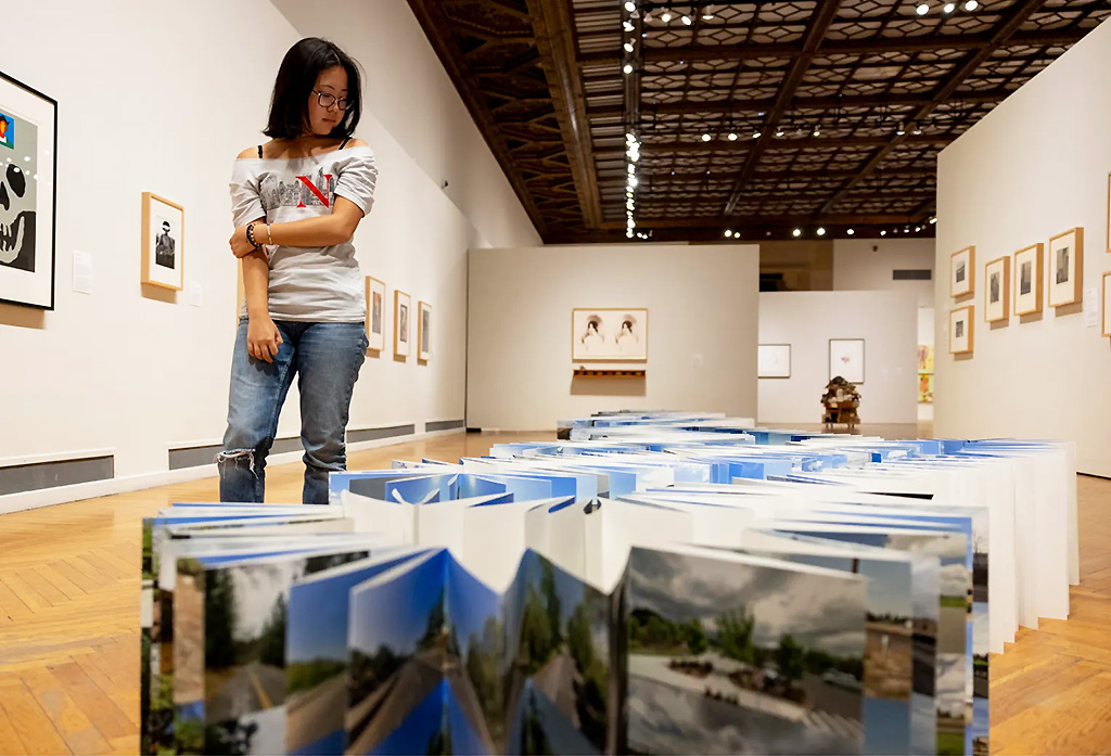 A woman in glasses and casual clothes looks at a large accordion-style open book displaying landscape photos on the floor of the Mills College Art Museum with framed artworks on the walls.