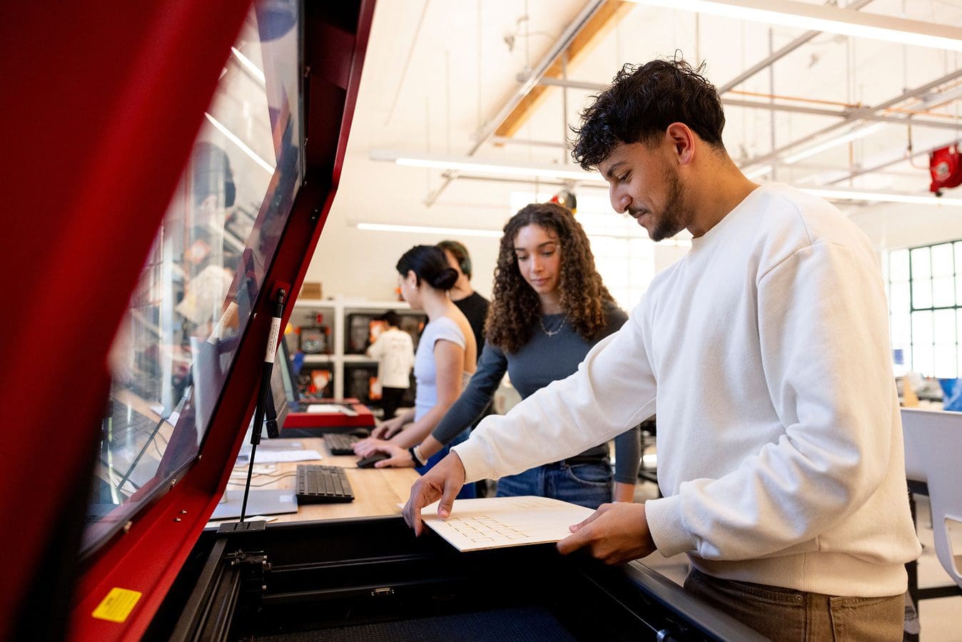 Makerspace gallery Three young adults work together in a modern lab space. One person operates a large red laser cutter, while two others use laptops at a nearby table. Bright, open room with shelves and equipment in the background.