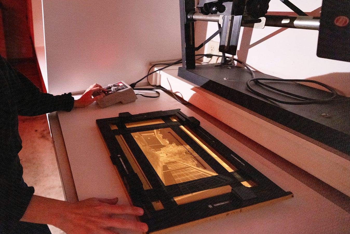 Makerspace gallery A person operates equipment in a darkroom, preparing to enlarge a photographic negative placed on an enlarger base under red safe lighting.