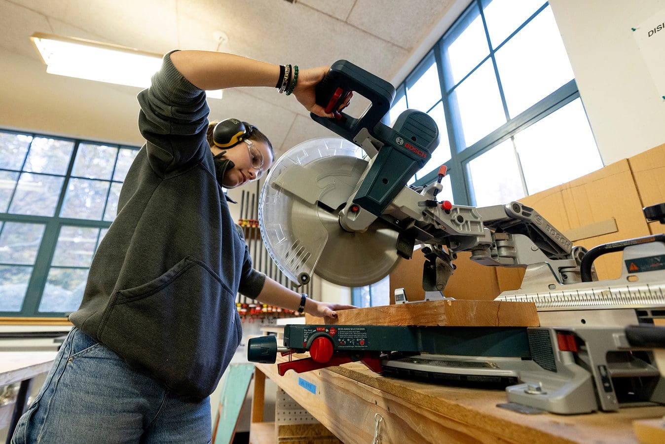 Makerspace gallery A person wearing safety goggles and earmuffs operates a circular miter saw to cut a piece of wood in a well-lit workshop with large windows.