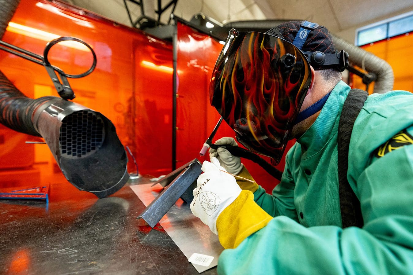 Makerspace gallery A person wearing a flame-pattern welding helmet and protective gloves works with metal sheets in a workshop, surrounded by safety equipment and orange plastic barriers.