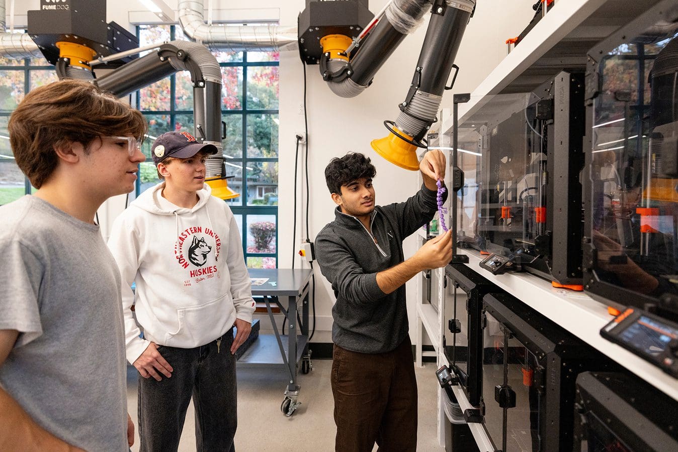Makerspace gallery Three young men in a lab observe as one holds up a 3D printed object in front of a row of 3D printers, with large ventilation hoses above and windows showing a view of trees outside.