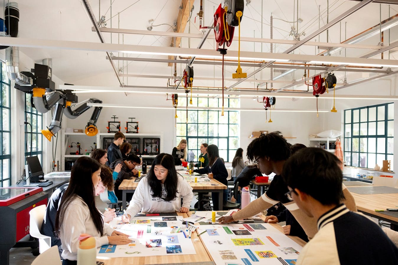 Makerspace gallery Students collaborate in a bright, modern classroom with large windows, using papers, laptops, and design materials. Some work in groups at tables, while others engage with equipment along the walls.