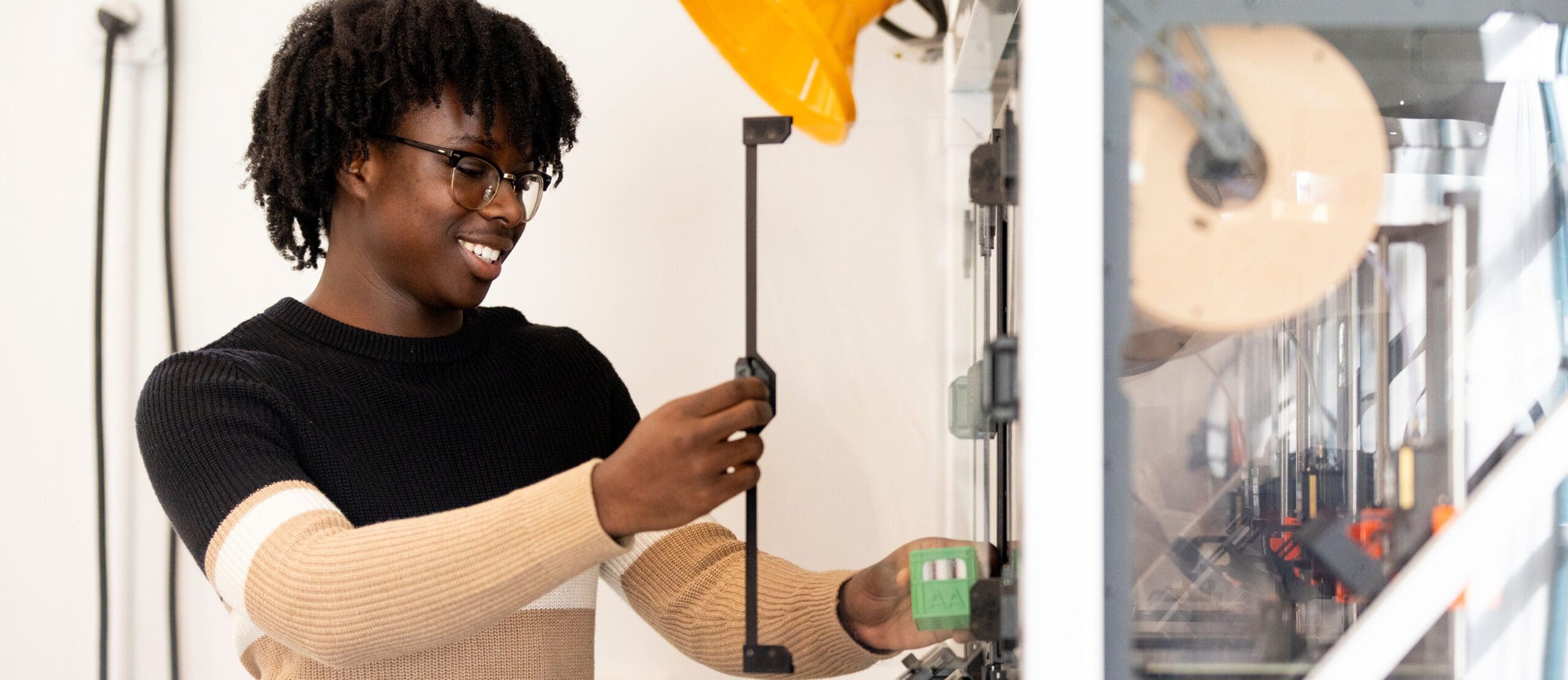 A person wearing glasses and a striped sweater smiles while using a 3D printer, holding a printed part and adjusting the printer inside a lab or workshop.