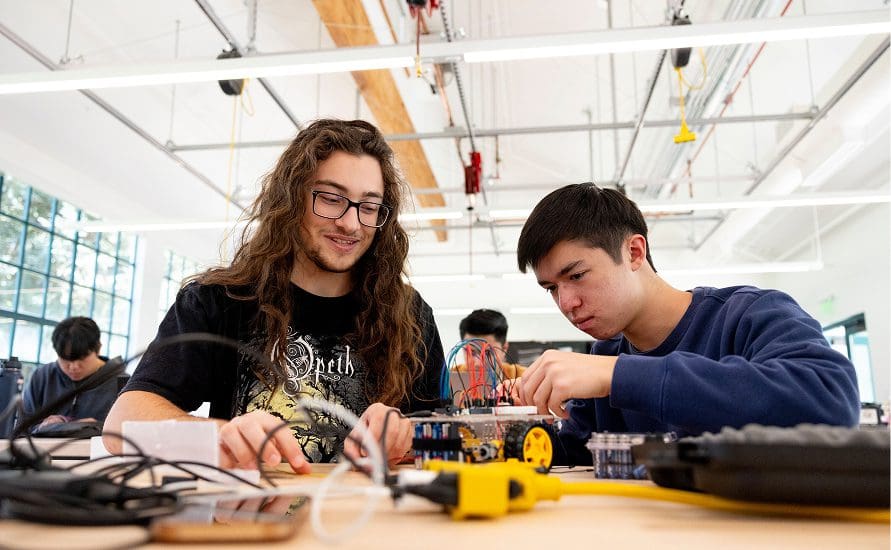 Two students work together on building a small robotic vehicle at a table in a bright, modern classroom filled with electronics and equipment.