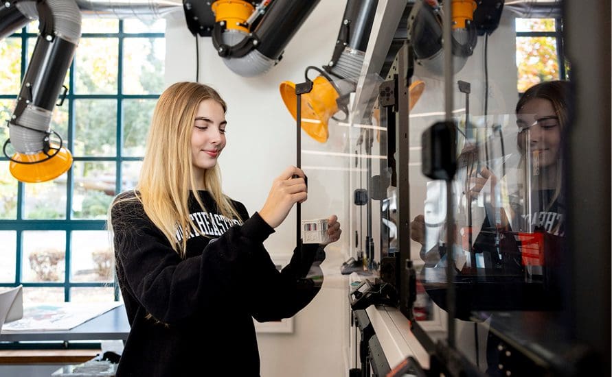 A young woman with long blonde hair uses a 3D printer in a bright, modern lab. She is wearing a black sweatshirt and holding a tool while operating the printer, with extraction vents and large windows in the background.