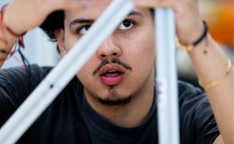 A person with a mustache and goatee looks intently at an object formed by three white bars, appearing focused and thoughtful, with hands reaching toward the structure.