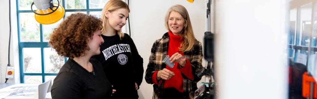Three people stand together indoors near a window. An older woman is explaining or showing something using a small device, while two younger women listen and watch attentively. Workshop tools are visible in the background.