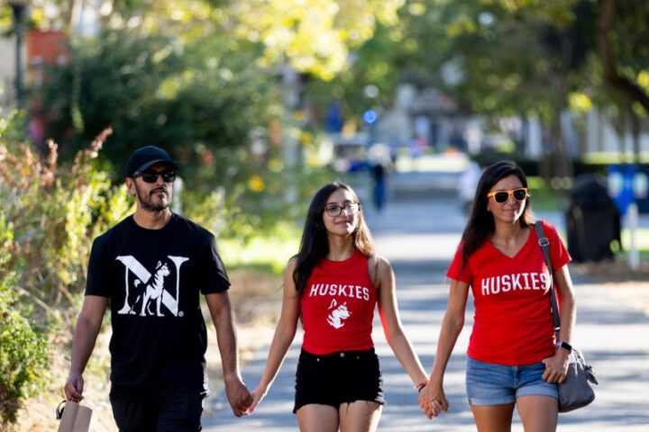 Three people wearing Husky spirit shirts walk up the path on Northeastern's Oakland campus along the Betty Irene Moore Natural Sciences Building.