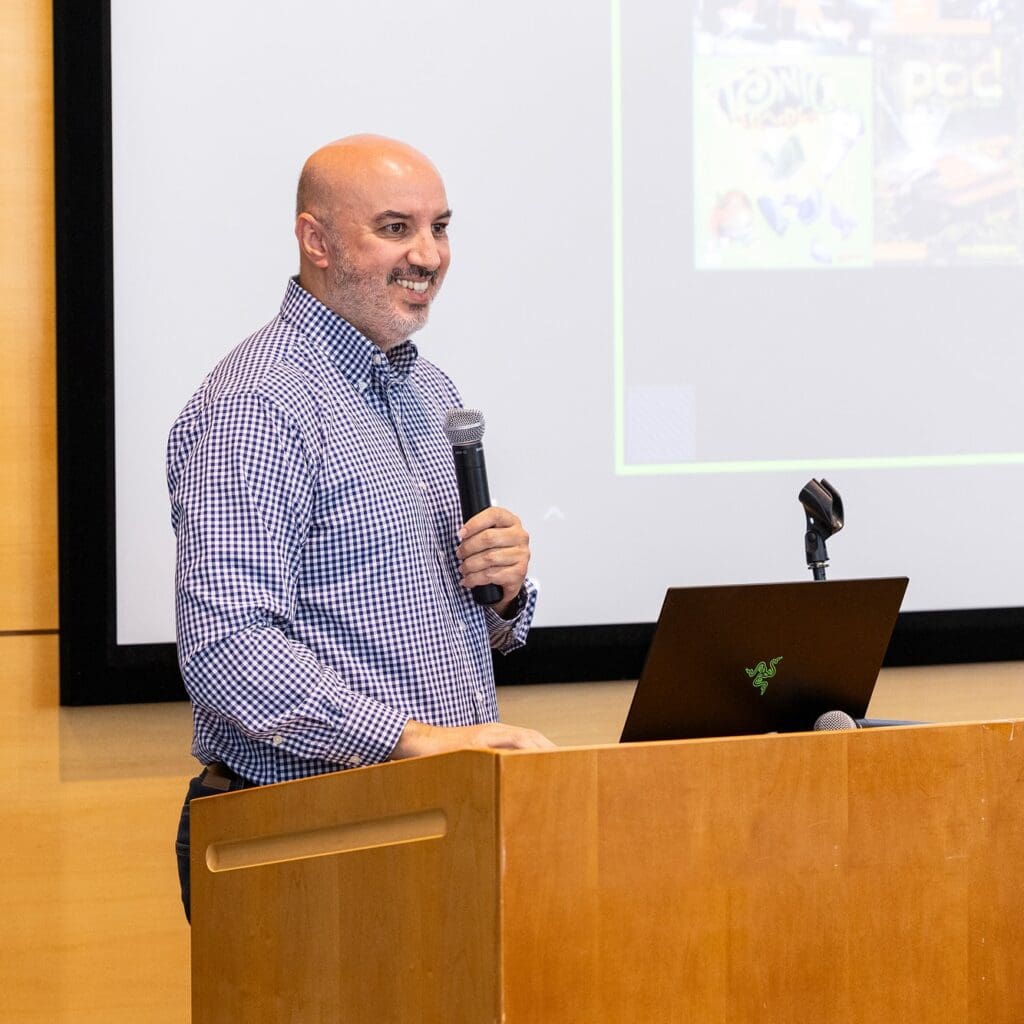 A man in a checkered shirt stands at a podium, holding a microphone and smiling, with a laptop in front of him and a presentation projected on a screen behind.