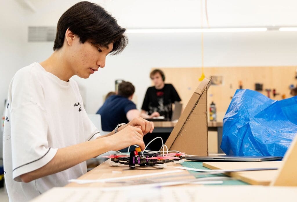 A young person in a white shirt works intently on assembling an electronic device at a table in a workshop, with tools and materials scattered around. Other people are seen working in the background.
