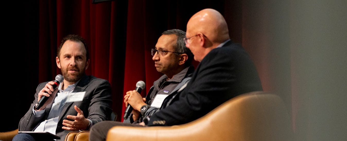 Three men sit in brown chairs on stage, engaged in a panel discussion. One man holds a microphone and gestures while speaking, while the other two listen attentively. Red curtains hang in the background.