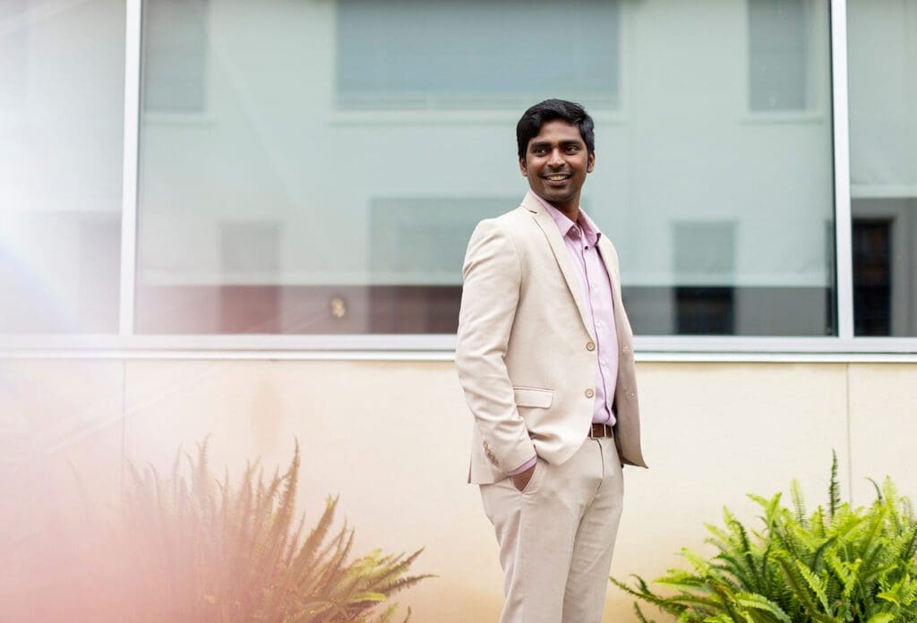A man in a light beige suit and pink shirt stands outdoors in front of a modern building with large windows, smiling and looking to his left. Green ferns are visible by his side.
