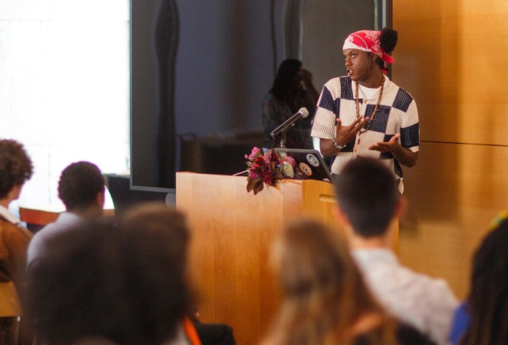 A person wearing a red bandana and patterned shirt speaks passionately at a podium with a microphone and laptop, addressing an audience in a well-lit room.