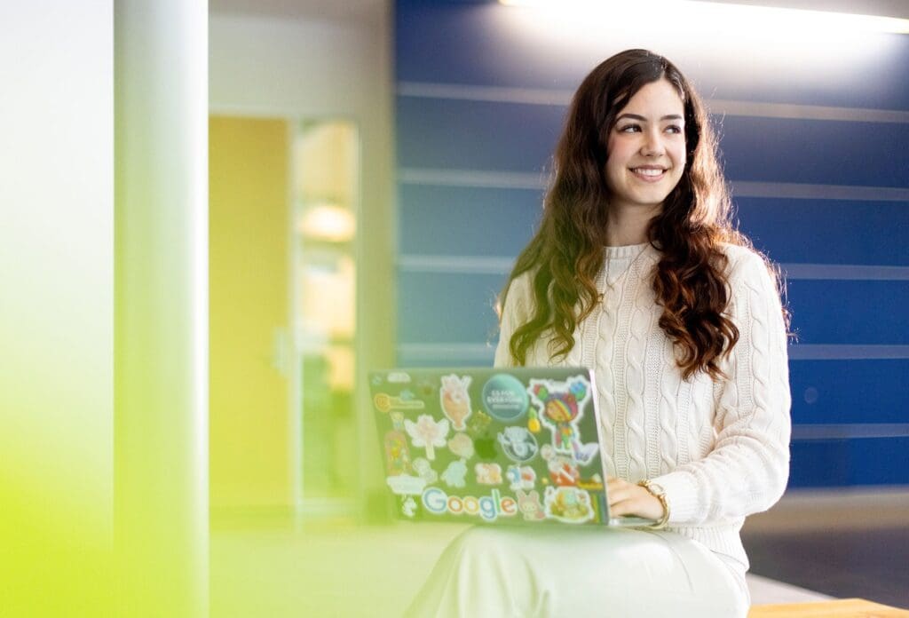 A woman with long dark hair, wearing a white sweater, sits and smiles while using a laptop decorated with colorful stickers, including one that says "Google," in a modern, blue-accented interior.