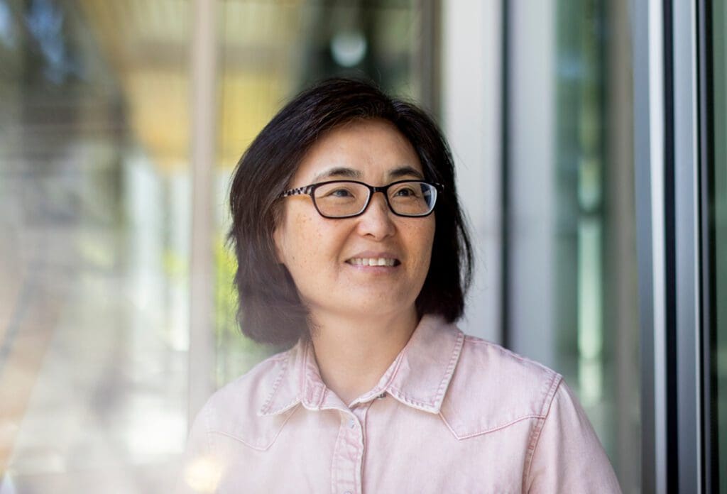 Miki Hong, wearing glasses and a light pink button-up shirt, smiles while looking slightly to the side. The background features glass doors and natural light.