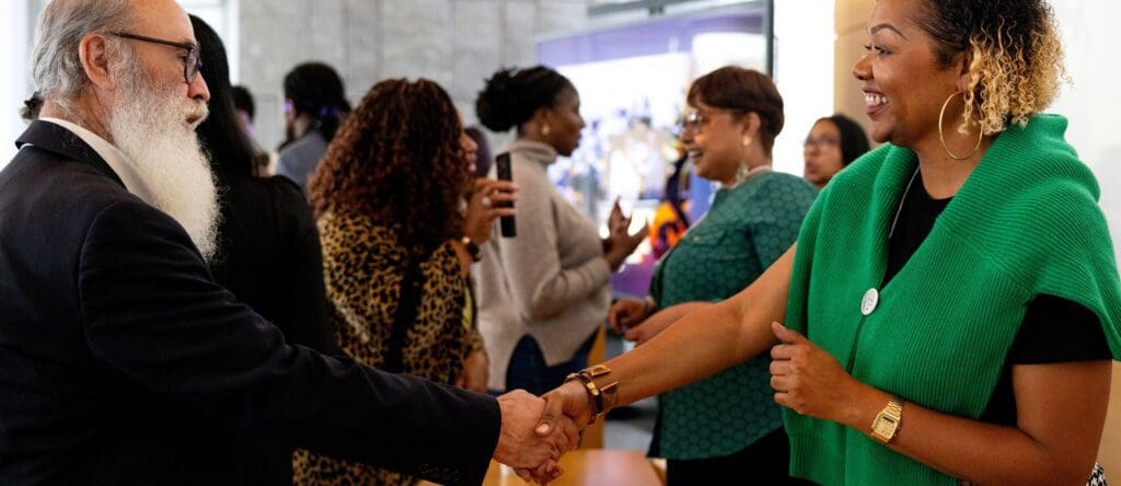 A man with a long white beard in a suit shakes hands with a smiling woman wearing a green sweater, as others mingle and talk in the background at a social or professional event.