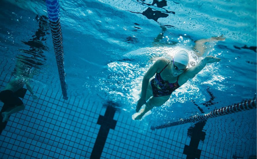 A swimmer wearing a white cap and goggles swims freestyle in a pool, viewed underwater. Lane ropes are visible on both sides, and another swimmer is seen in a neighboring lane.