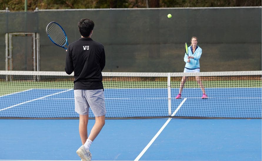 Two people play tennis on an outdoor court; one person in a black jacket and gray shorts prepares to hit the ball, while the other, in a light blue jacket, stands ready on the opposite side of the net.