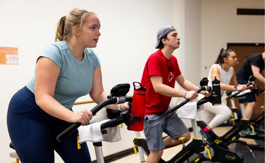 Three people exercise on stationary bikes in a gym. A woman in a blue top and navy leggings is in the foreground, while a man in a red shirt and a woman in white workout clothes ride bikes nearby.