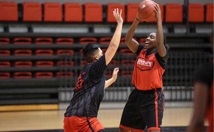 Two basketball players practice on an indoor court. One player in a red "Huskies" jersey holds the ball overhead while being closely guarded by a teammate wearing black and red. The bleachers are empty in the background.