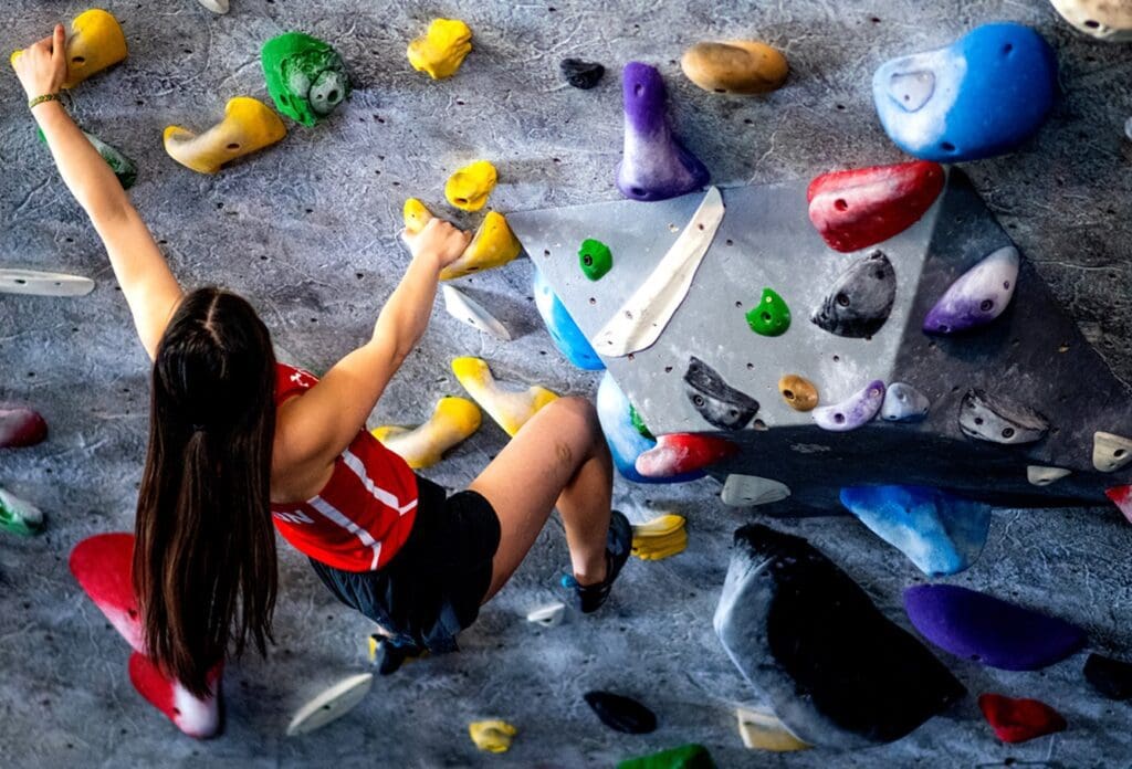 A woman in a red tank top and shorts climbs a colorful indoor bouldering wall, gripping holds of various colors and shapes as she reaches upward with one arm.