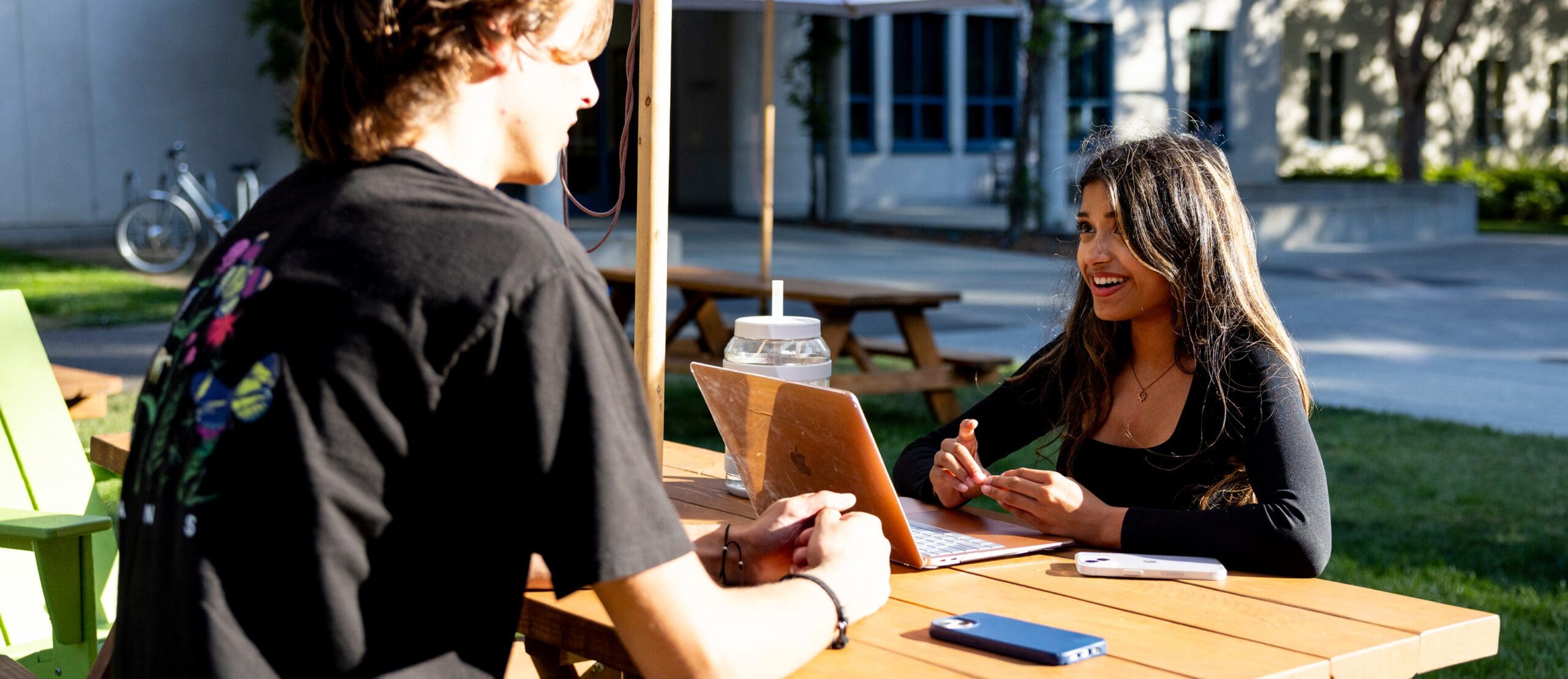 Two people sit at a wooden table outdoors. One is using a laptop and smiling, while the other listens. There are benches, a cup with a straw, a phone, and a notebook on the table. Bright sunlight and greenery are in the background.