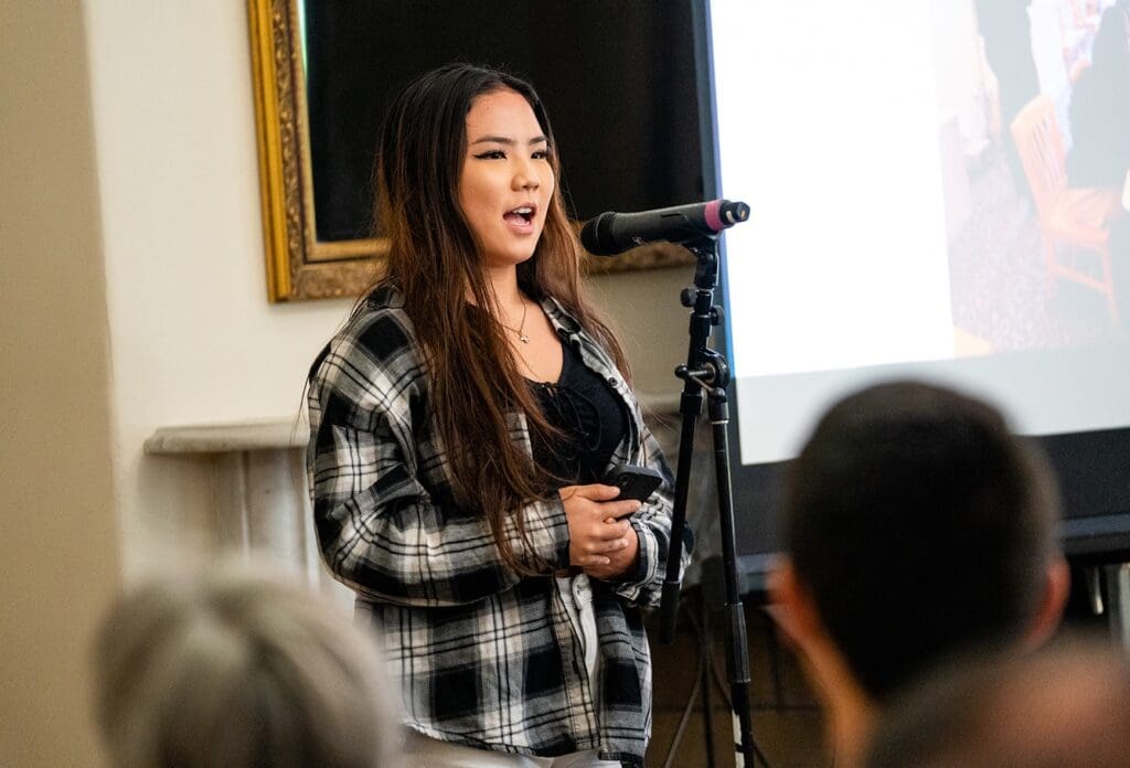 A young woman with long hair speaks into a microphone while standing in front of an audience. She wears a plaid shirt over a black top, and a large screen and a painting are visible behind her.