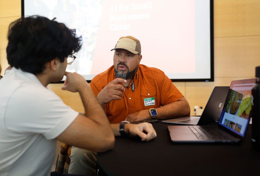 Two men sit at a table with laptops, having a focused conversation. One man, wearing an orange shirt and beige cap, gestures while speaking. A presentation screen behind them reads "AI for Small Businesses Clinic.
