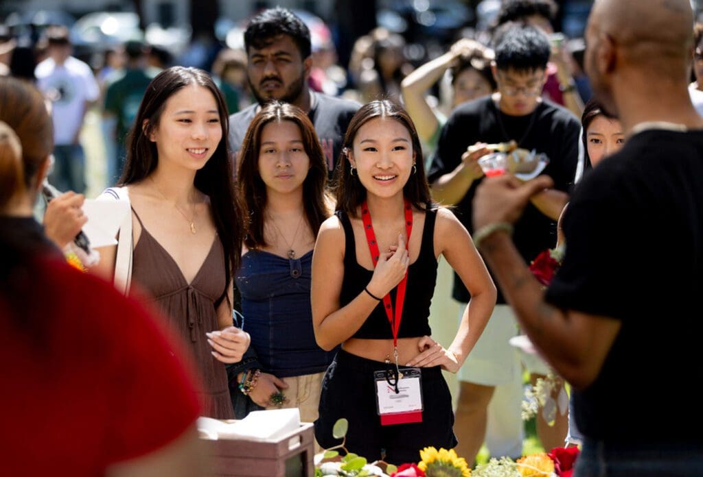 A group of young adults, including three women in front, stand smiling at an outdoor table with flowers and food, surrounded by other people in a sunny, busy setting.