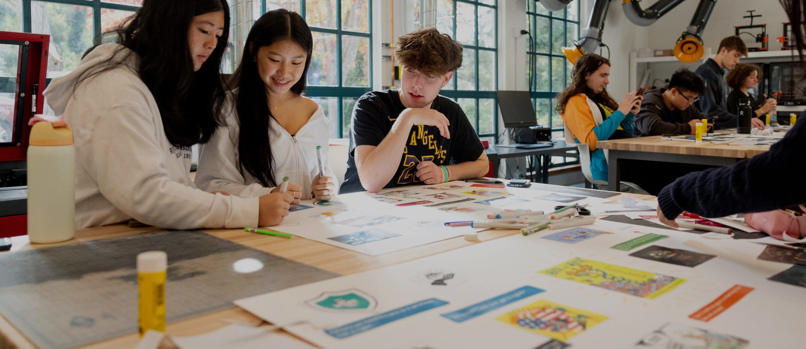 Students work together at a table filled with art supplies and colorful papers in a bright classroom, while others collaborate in the background.