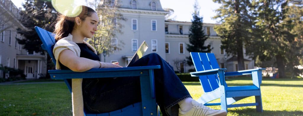 A young woman sits on a bright blue outdoor chair on a sunny lawn, working on a laptop, with historic campus buildings and trees in the background.