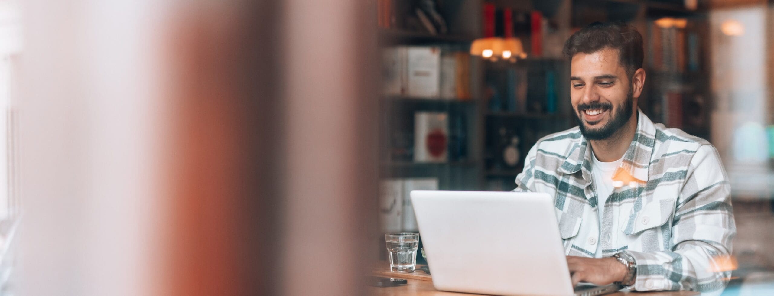 A man wearing a plaid shirt sits at a table in a cozy café, smiling while working on a laptop. A glass of water is next to him, and bookshelves are visible in the blurred background.