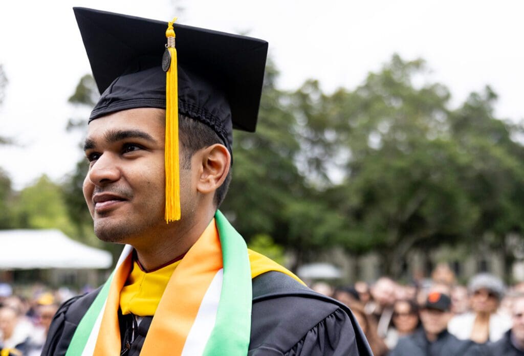 A young graduate in a cap and gown smiles outdoors, wearing a colorful sash, with trees and a blurred crowd in the background.
