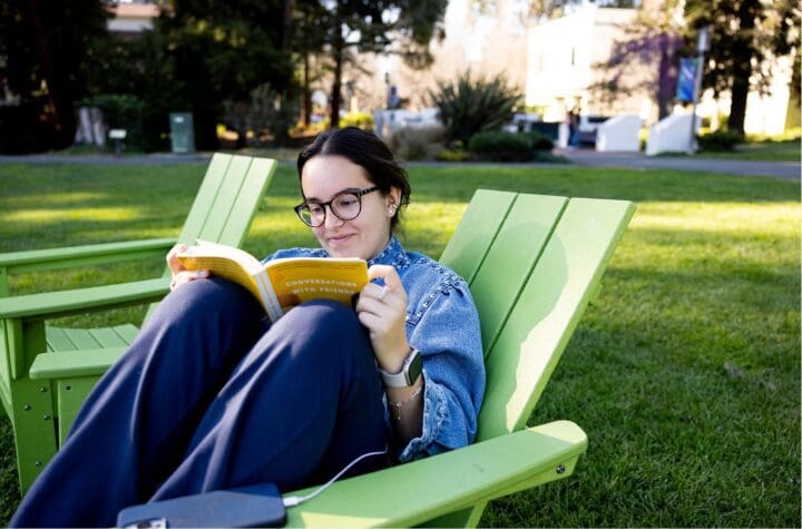 Student sits in lawn chair reading behind Mills Hall