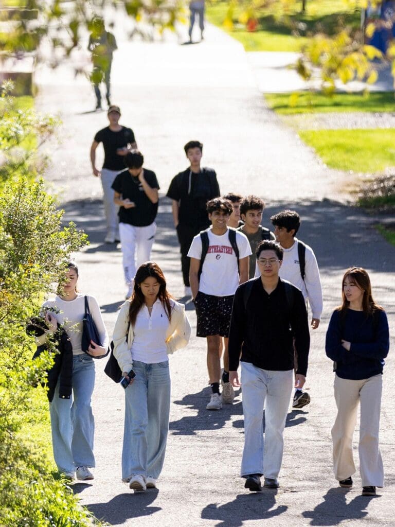 A group of students walk along a sunlit campus pathway, some chatting in pairs while others walk alone. Green grass and trees border the path, and the students appear relaxed in casual clothing.