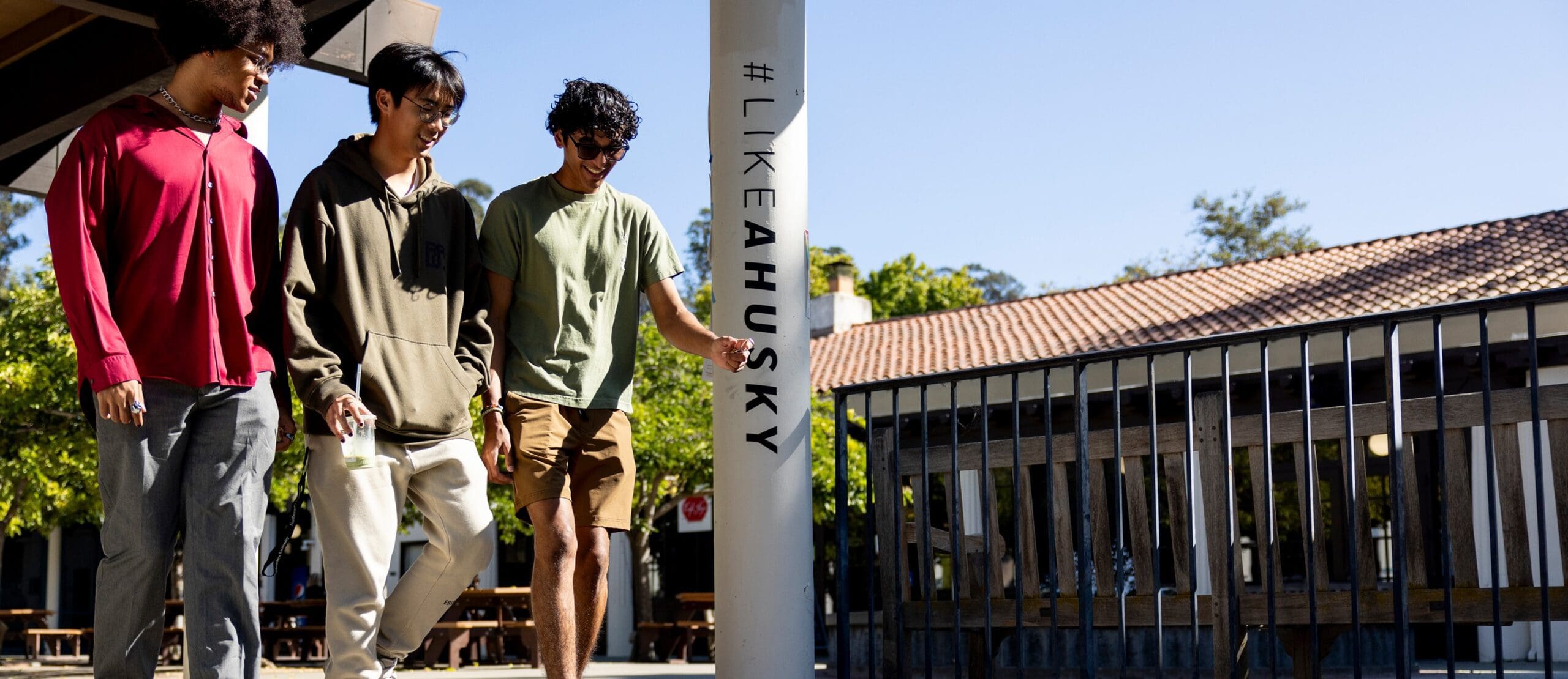 Three young men walk together outdoors near a white pole labeled "#LIKEAHUSKY," with trees and a building in the background on a sunny day.