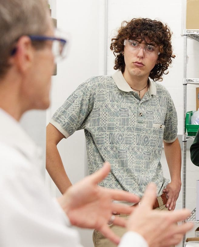 A young person with curly hair and safety glasses listens attentively to someone speaking, who is out of focus in the foreground. They are standing in a room with white walls and metal shelves.