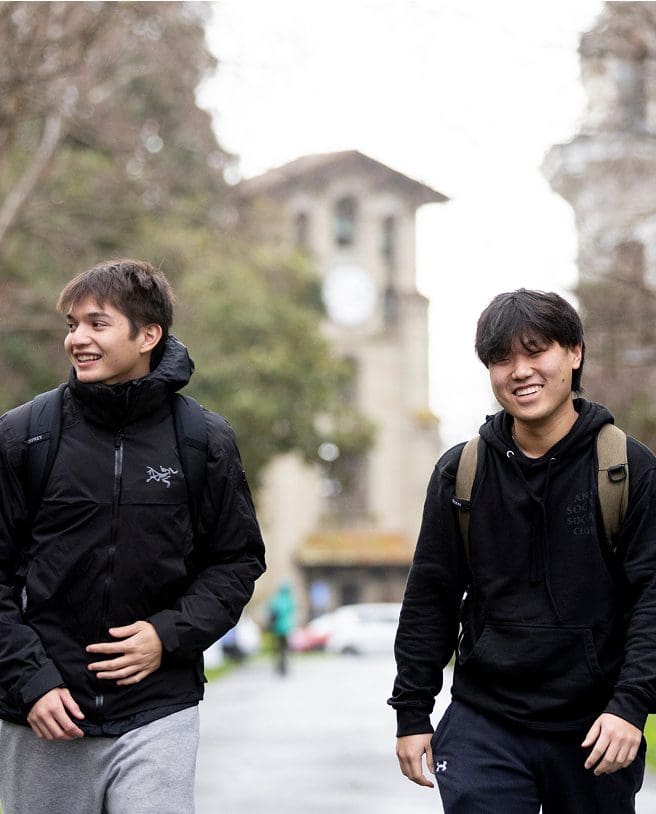 Two young men wearing black jackets and backpacks walk outdoors, smiling. Trees and a building with a clock tower appear blurred in the background. The weather looks cool and overcast.