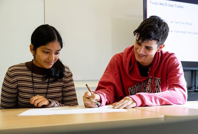 Two students sit at a desk working together on a sheet of paper. One student is writing while the other looks on. A presentation screen is visible in the background.