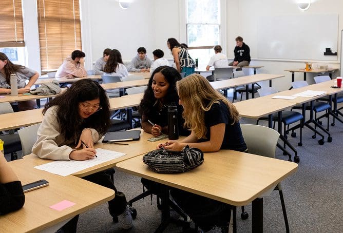 Students sit in groups at tables in a classroom, working together on assignments. Three students in the foreground are engaged in discussion and writing, while others collaborate in the background.