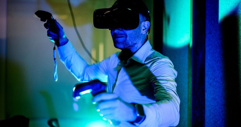 A man wearing a virtual reality headset and holding motion controllers in both hands stands in a room with blue and green lighting, fully immersed in the VR experience.