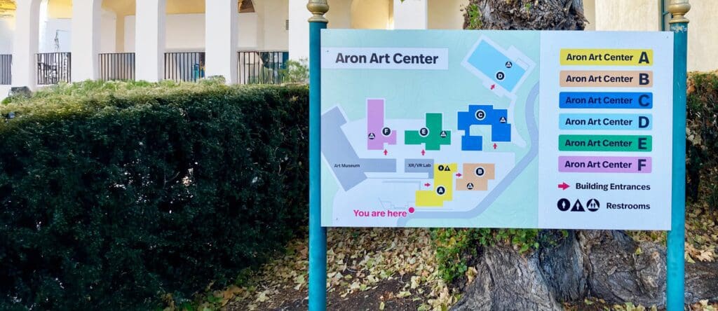 A sign displays a colorful map of the Aron Art Center, marking buildings A–F, entrances, and restrooms. The map is outdoors, near bushes and a tree, with part of a building visible in the background.