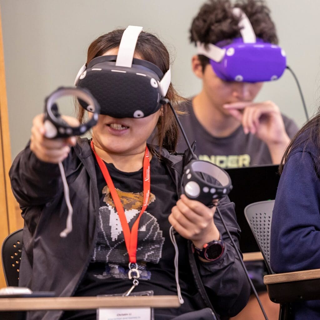 A person wearing a virtual reality headset and holding controllers is engaged in a VR experience, while another person in the background also wears a headset and looks down thoughtfully.