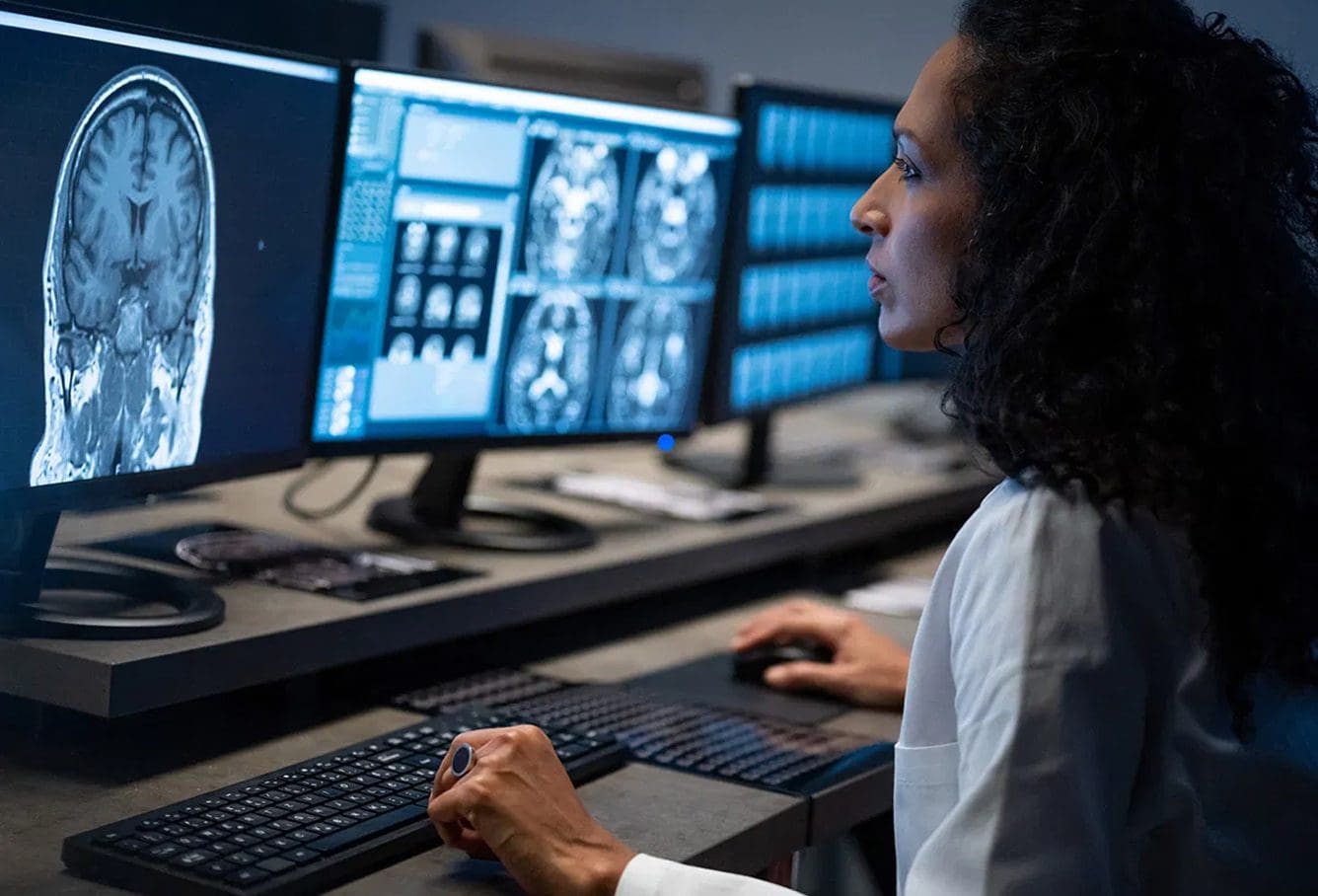 A woman in a lab coat analyzes brain scans on multiple computer monitors in a dimly lit control room, using a keyboard and mouse.